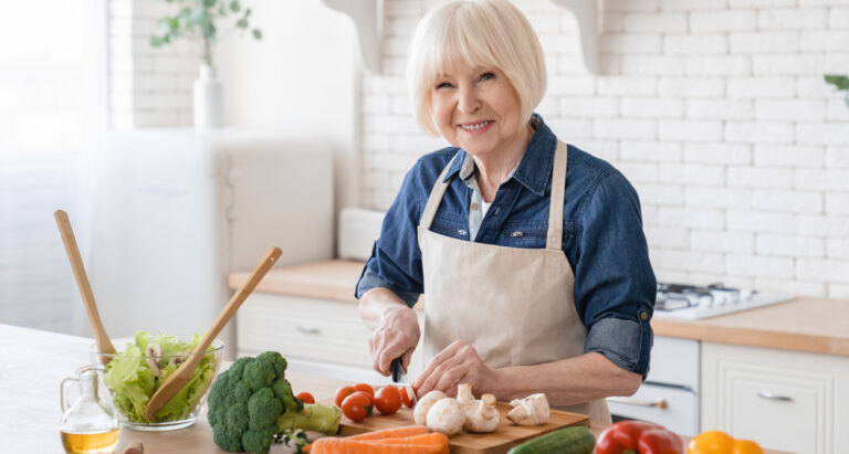 A senior woman chopping tomatoes on a cutting board in her kitchen with other vegetables on the counter while she smiles