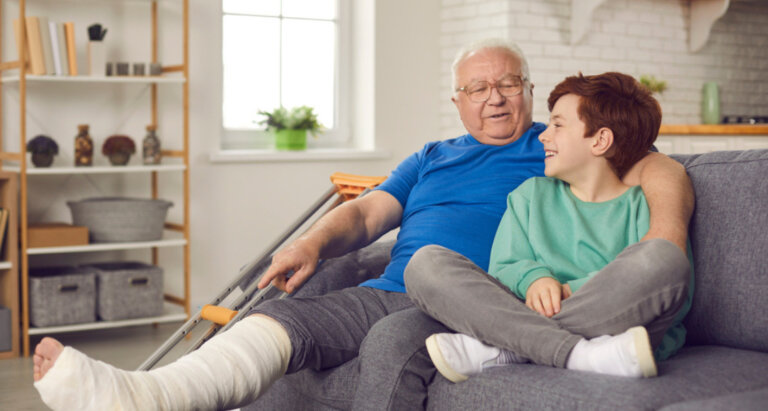 A senior man sitting on a grey couch with his arm around his grandson's shoulder as he props his leg up with it in a cast